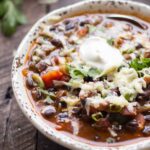 A close-up of a single bowl of black bean soup topped with sour cream, melted cheese, and chopped cilantro.