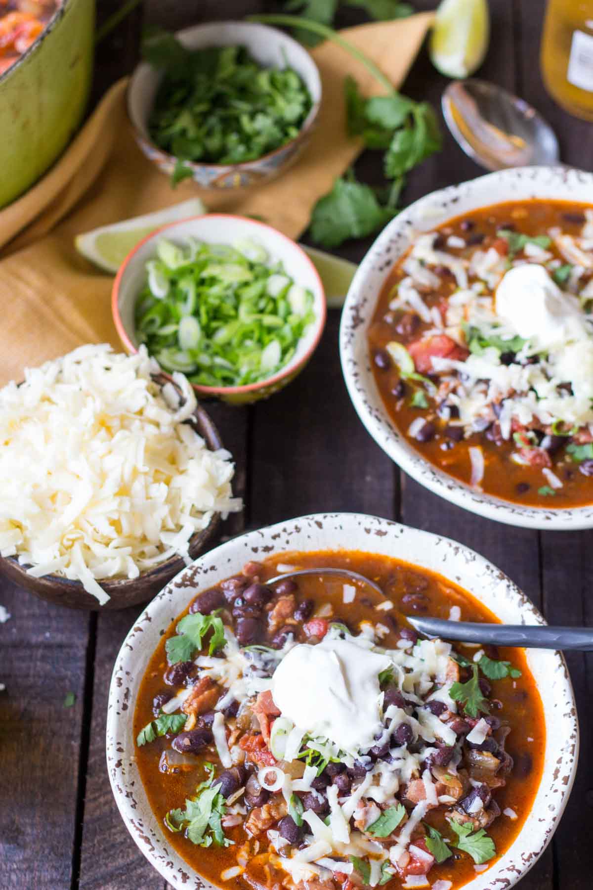 Overhead shot of two bowls of black bean soup garnished with sour cream, shredded cheese, and cilantro, with bowls of green onions and cheese arranged on the side.