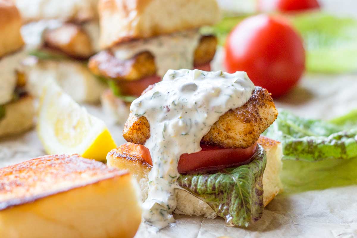 Close-up of a toasted slider bun filled with a piece of blackened fish, fresh tomato, crisp lettuce, and a generous drizzle of creamy tartar sauce, with a lemon wedge in the background.