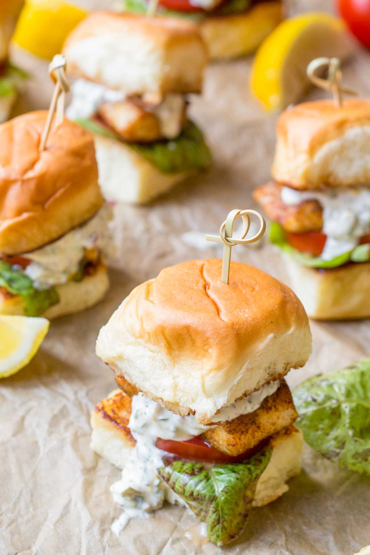 Overhead view of several blackened fish sliders on parchment paper, each topped with lettuce, tomato, and tartar sauce, surrounded by lemon wedges.
