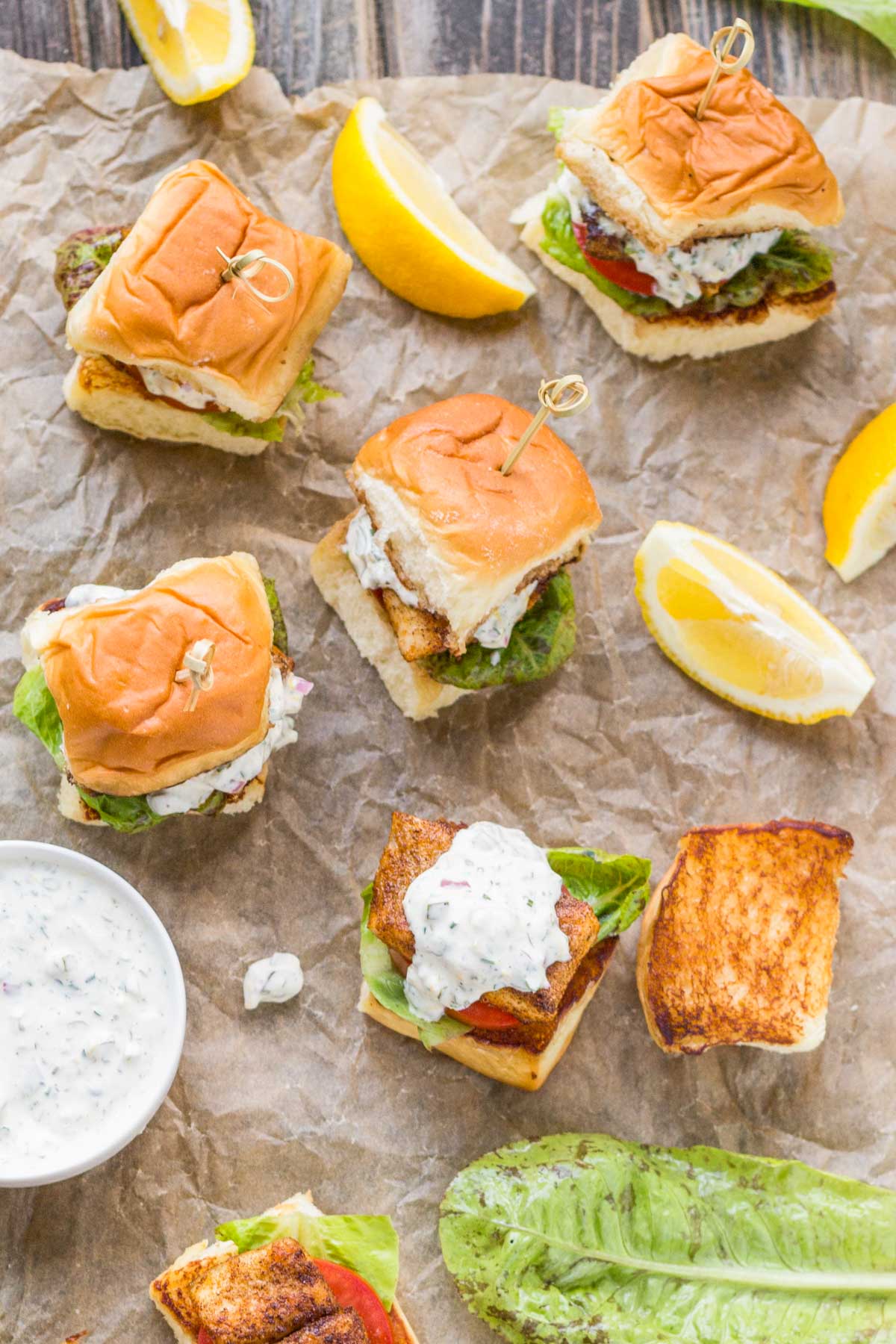 Assorted blackened fish sliders arranged on crinkled parchment with lemon wedges, showing toasted tops of the buns and creamy sauce spilling over the fish.