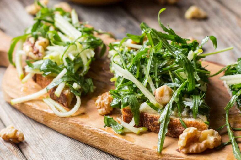 Close-up of apple tartines topped with arugula, walnuts, and julienned apples on toasted bread, served on a wooden board.