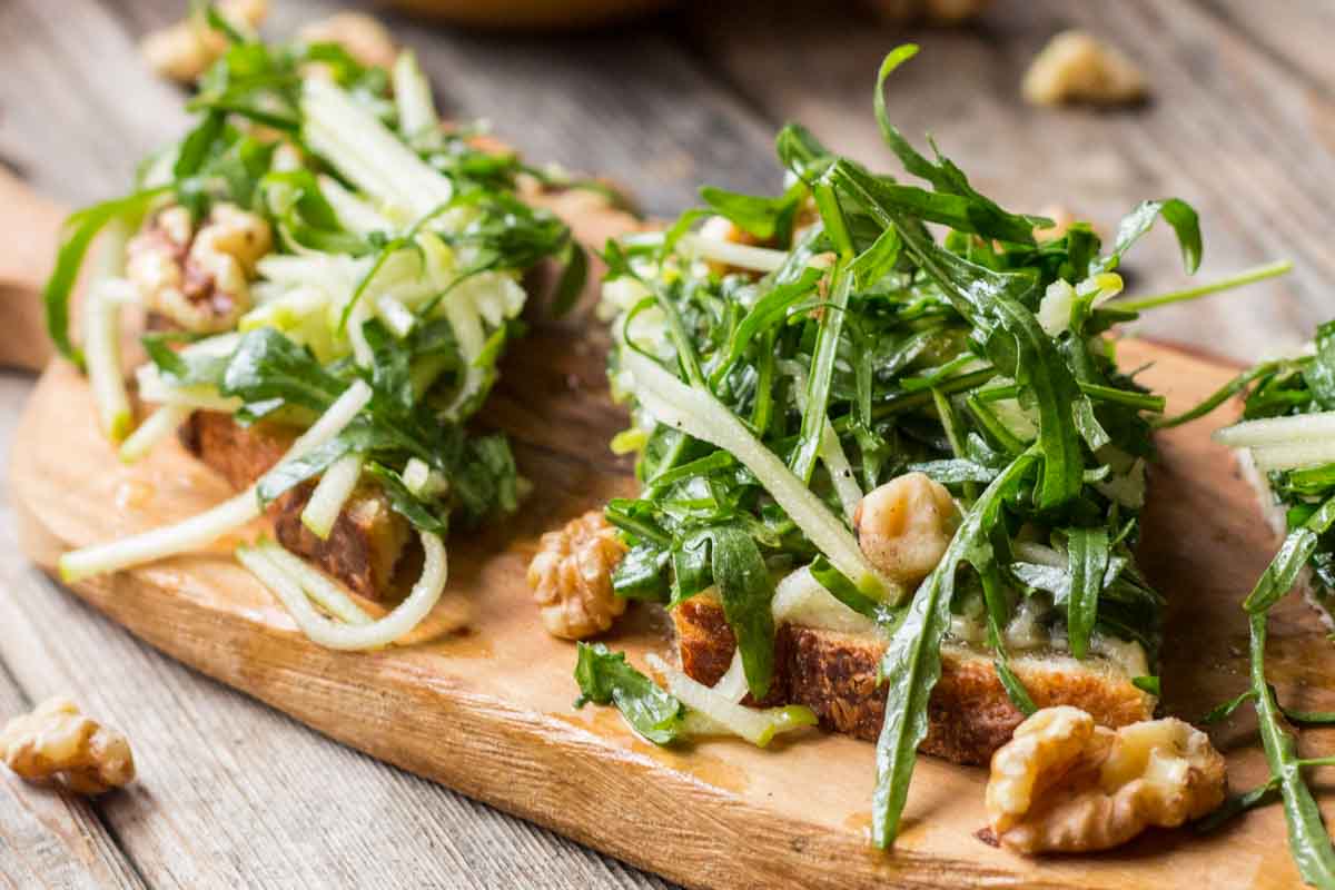 Close-up of apple tartines topped with arugula, walnuts, and julienned apples on toasted bread, served on a wooden board.