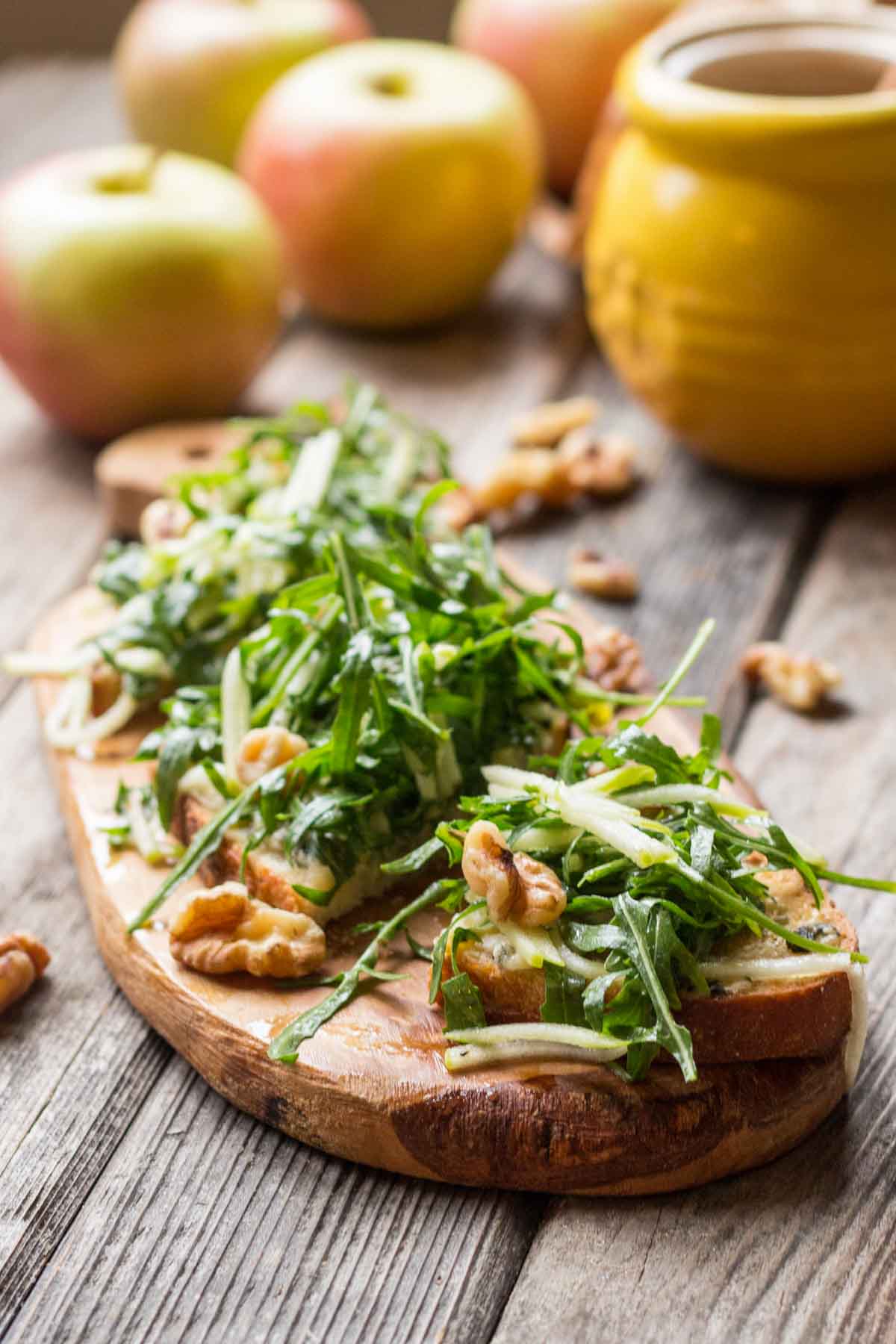 Apple tartines with arugula and walnuts arranged on a rustic wooden board, with fresh apples and a yellow honey pot in the background.