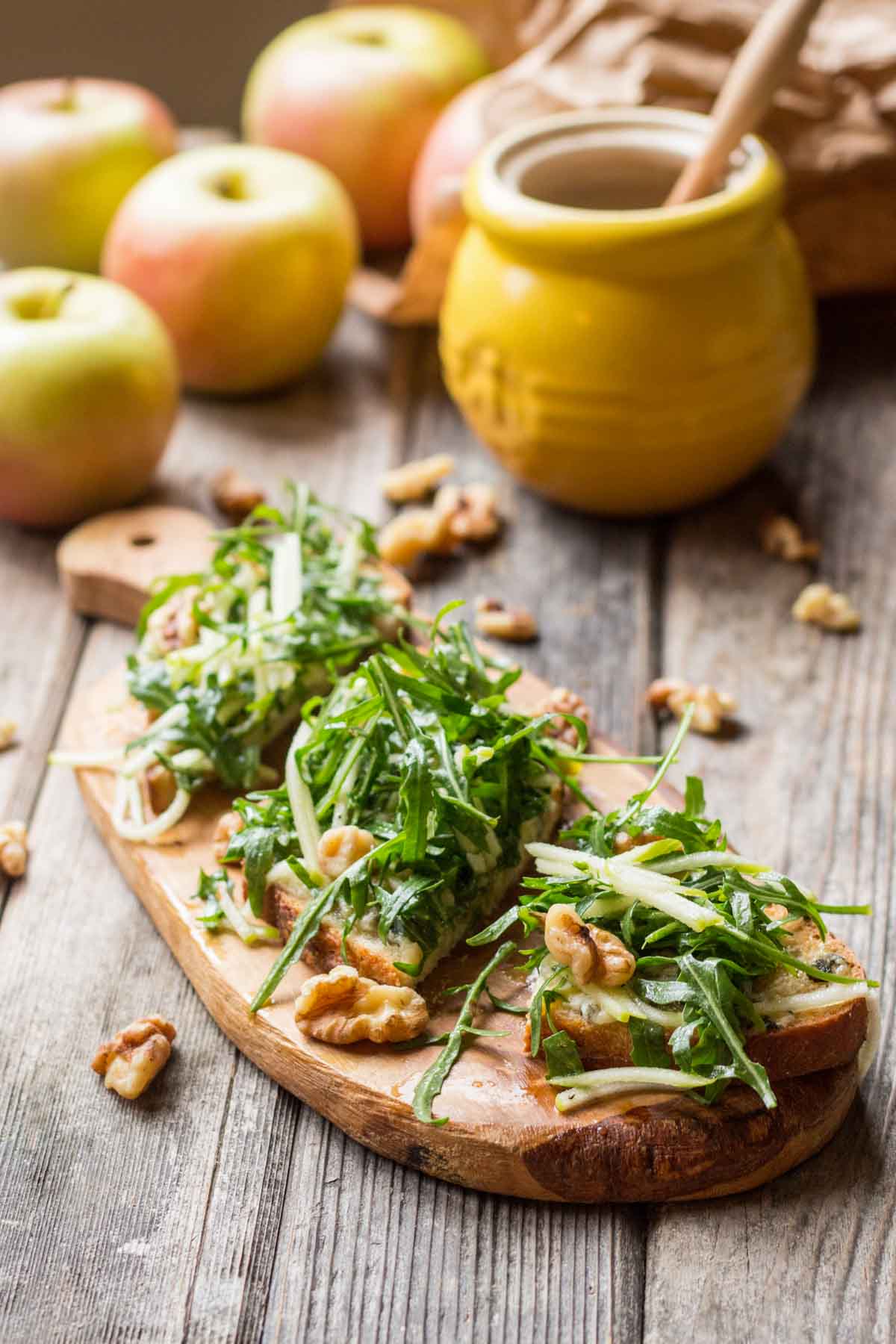 Three apple tartines layered with blue cheese, arugula, walnuts, and julienned apples on toasted bread, set on a wooden board with scattered walnuts.