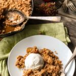 A white plate with a serving of brown butter apple crisp topped with a scoop of vanilla ice cream, set on a rustic wooden table with the skillet in the background.