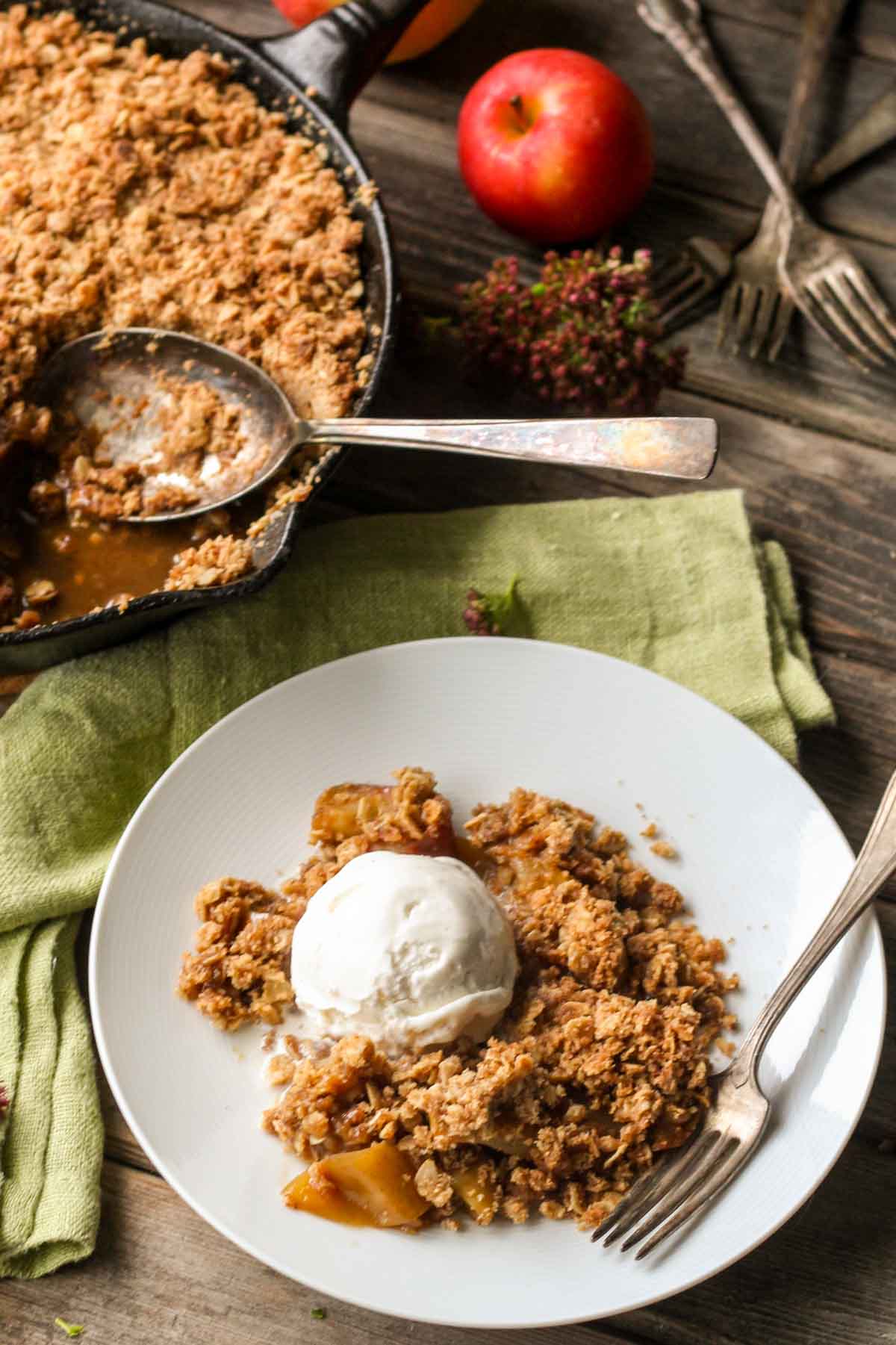 A white plate with a serving of brown butter apple crisp topped with a scoop of vanilla ice cream, set on a rustic wooden table with the skillet in the background.