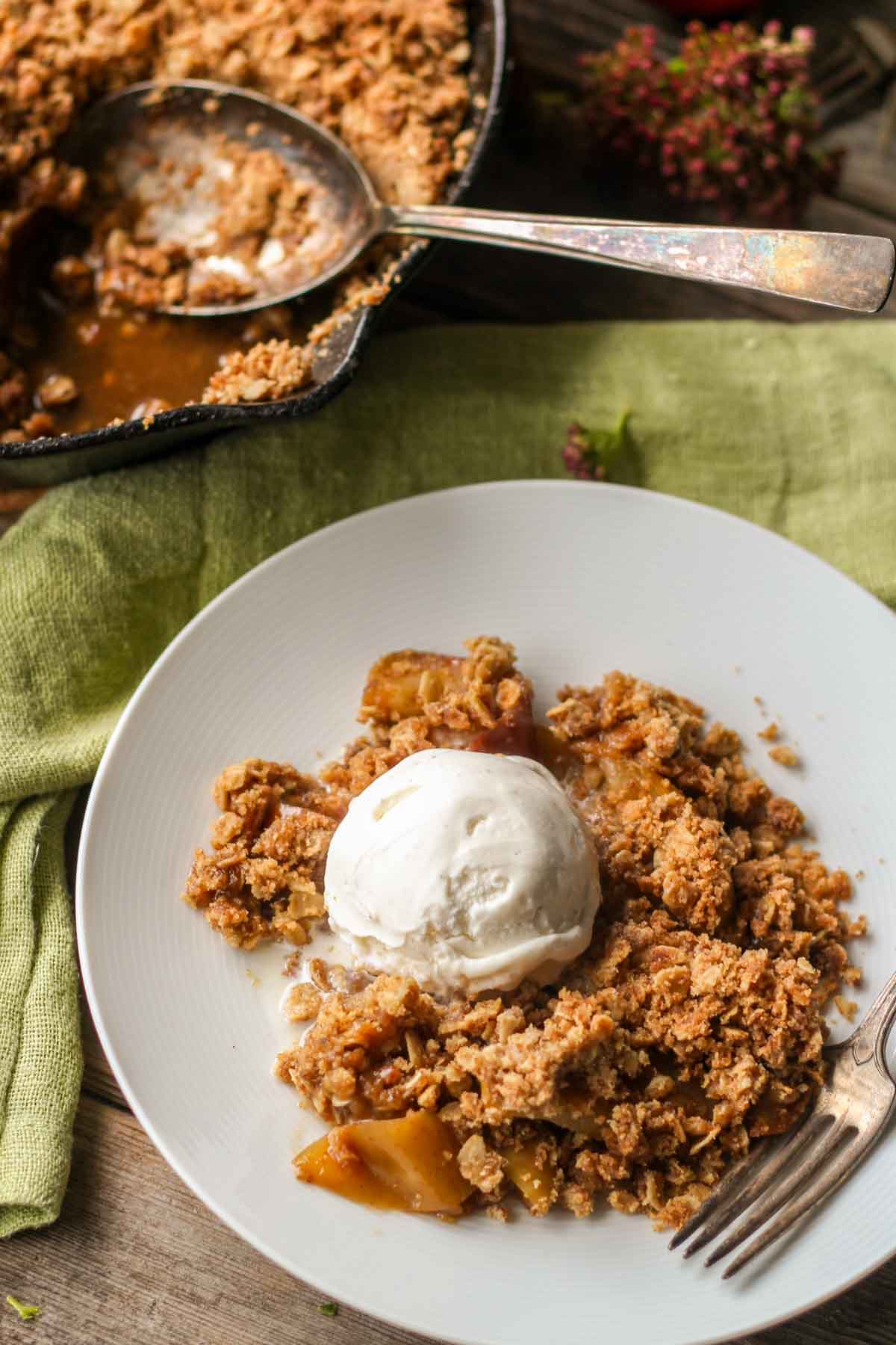 Rustic scene with a plate of brown butter apple crisp and vanilla ice cream in the foreground, with the skillet of crisp and serving spoon in the background.