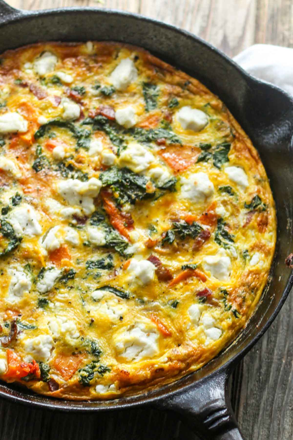 Close-up of butternut squash frittata in a cast iron skillet - Golden baked eggs filled with kale, orange squash, bacon, and dollops of white goat cheese.