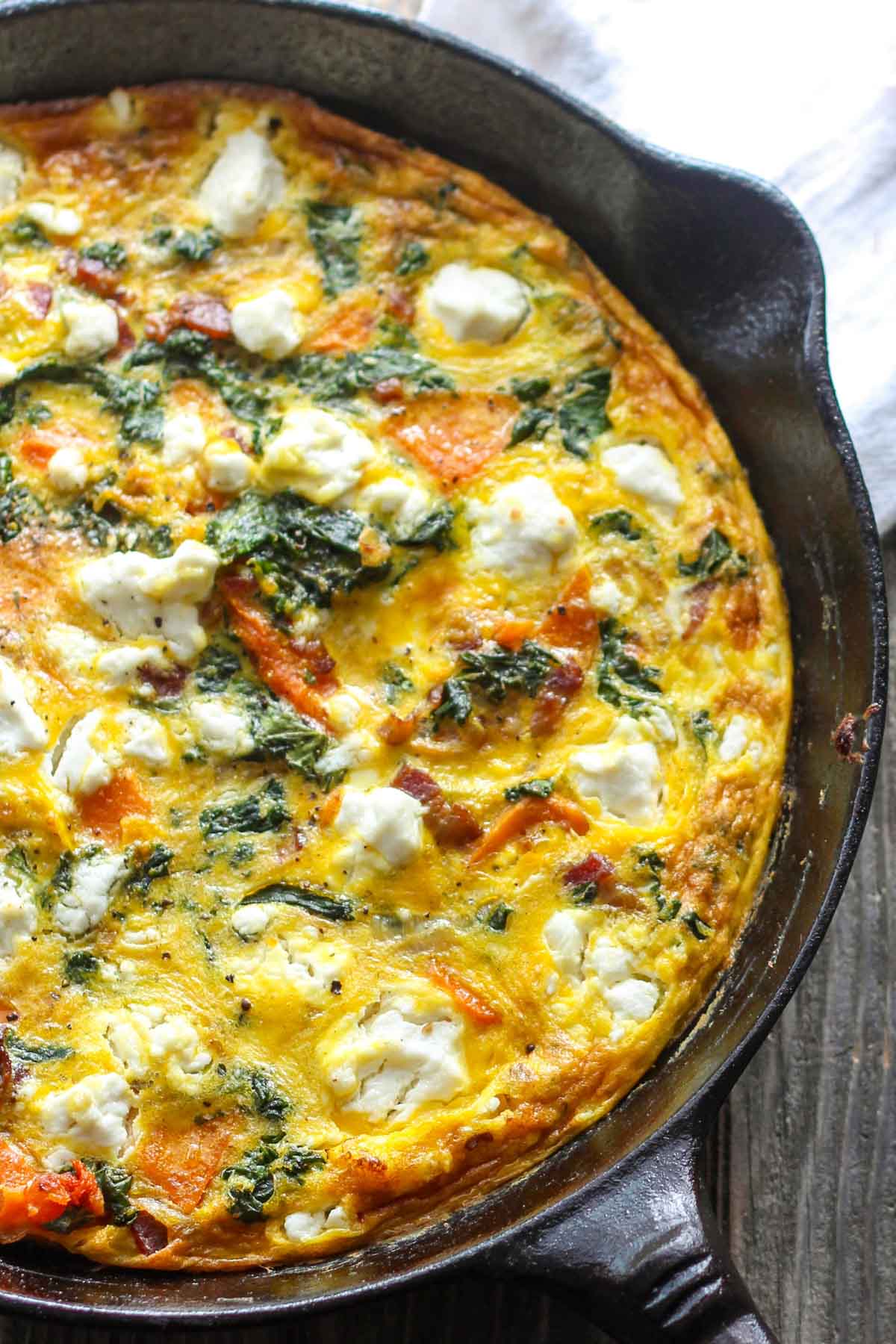 Side angle of baked frittata in cast iron skillet - Colorful chunks of orange squash and green kale set in fluffy golden eggs with goat cheese.