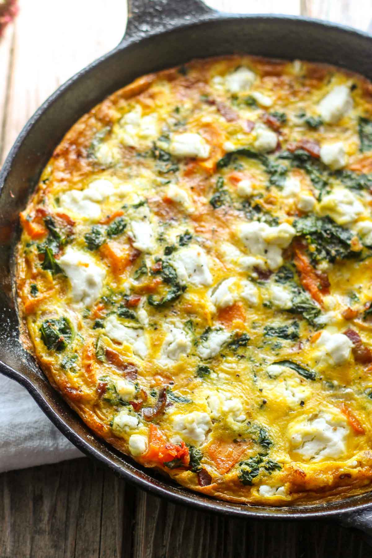 Butternut squash frittata in skillet on wooden table - Lightly browned edges with visible kale, bacon, and creamy goat cheese throughout.
