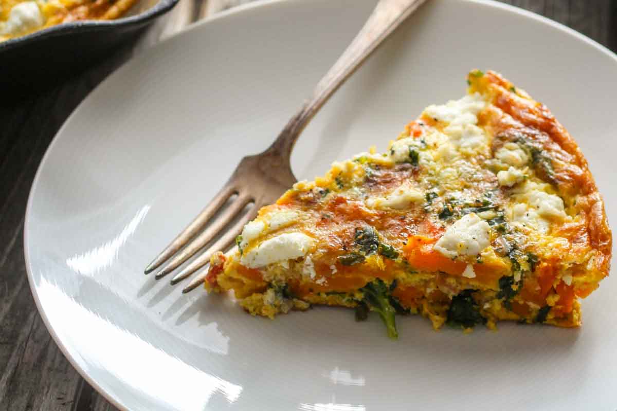 Slice of butternut squash frittata on white plate - Served with a fork, showing layers of orange squash, green kale, and white goat cheese in golden eggs.