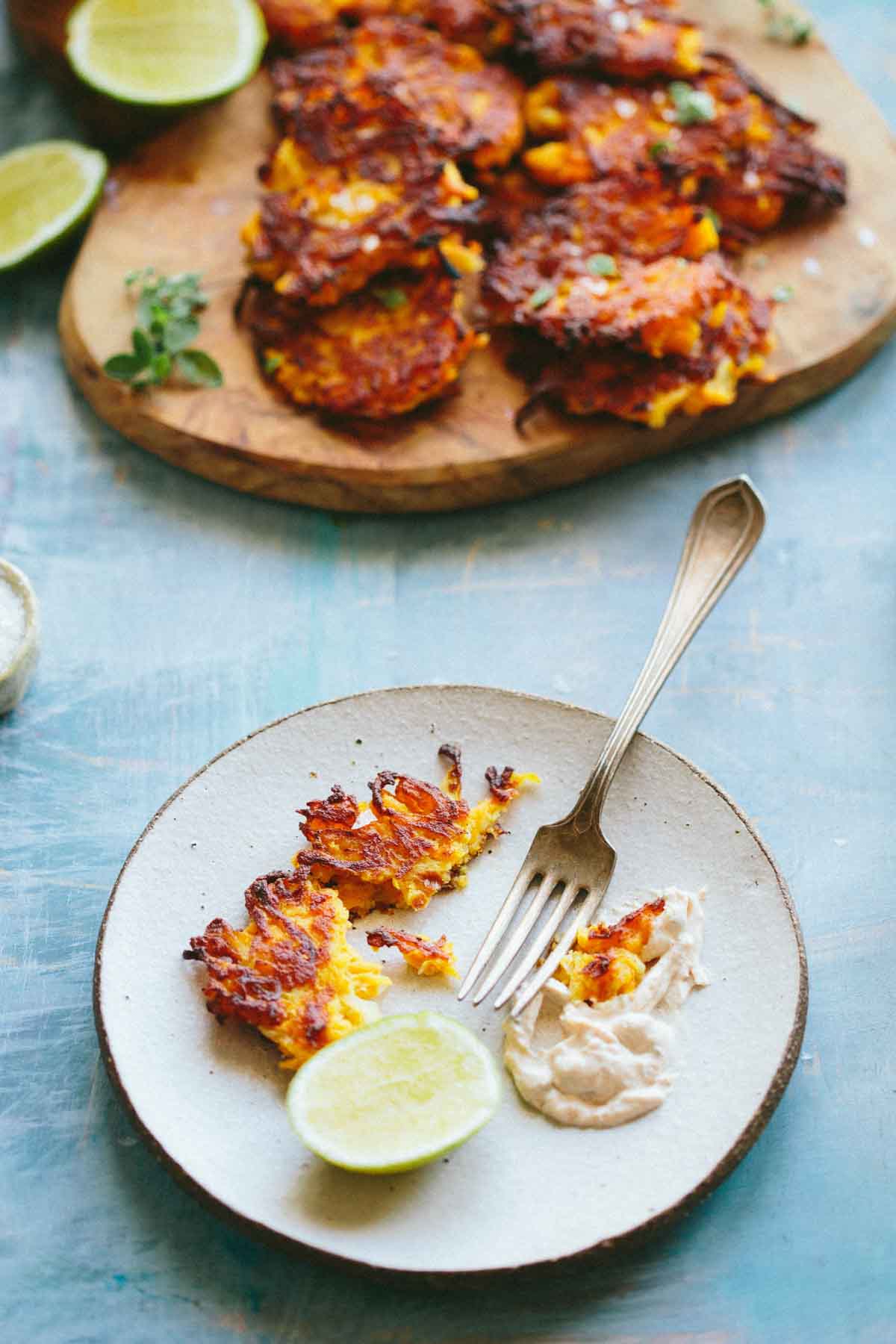 A plate with broken pieces of fritters, lime wedges, and spiced yogurt for dipping, with more fritters piled on a wooden board in the background.