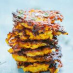 A tall stack of crispy butternut fritters glistening with olive oil, garnished with small green herbs against a light blue surface.