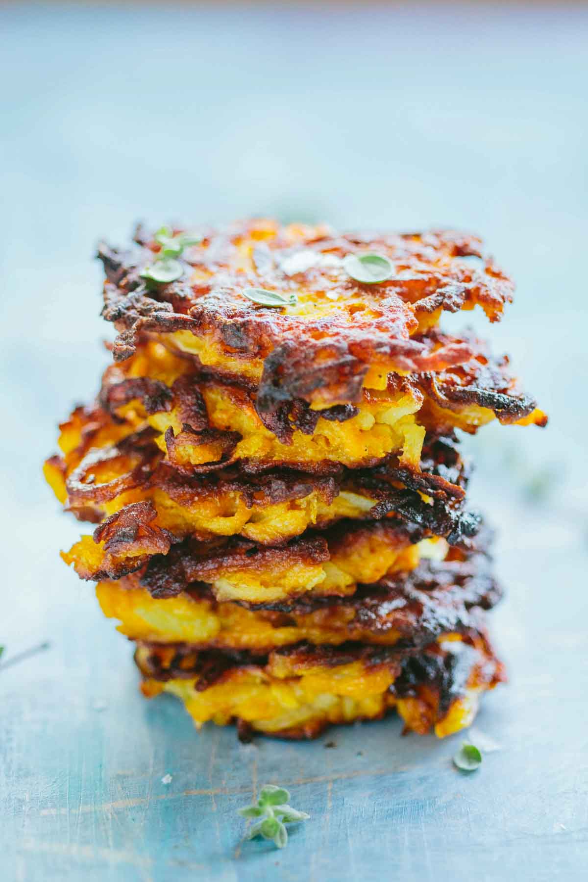A tall stack of crispy butternut fritters glistening with olive oil, garnished with small green herbs against a light blue surface.