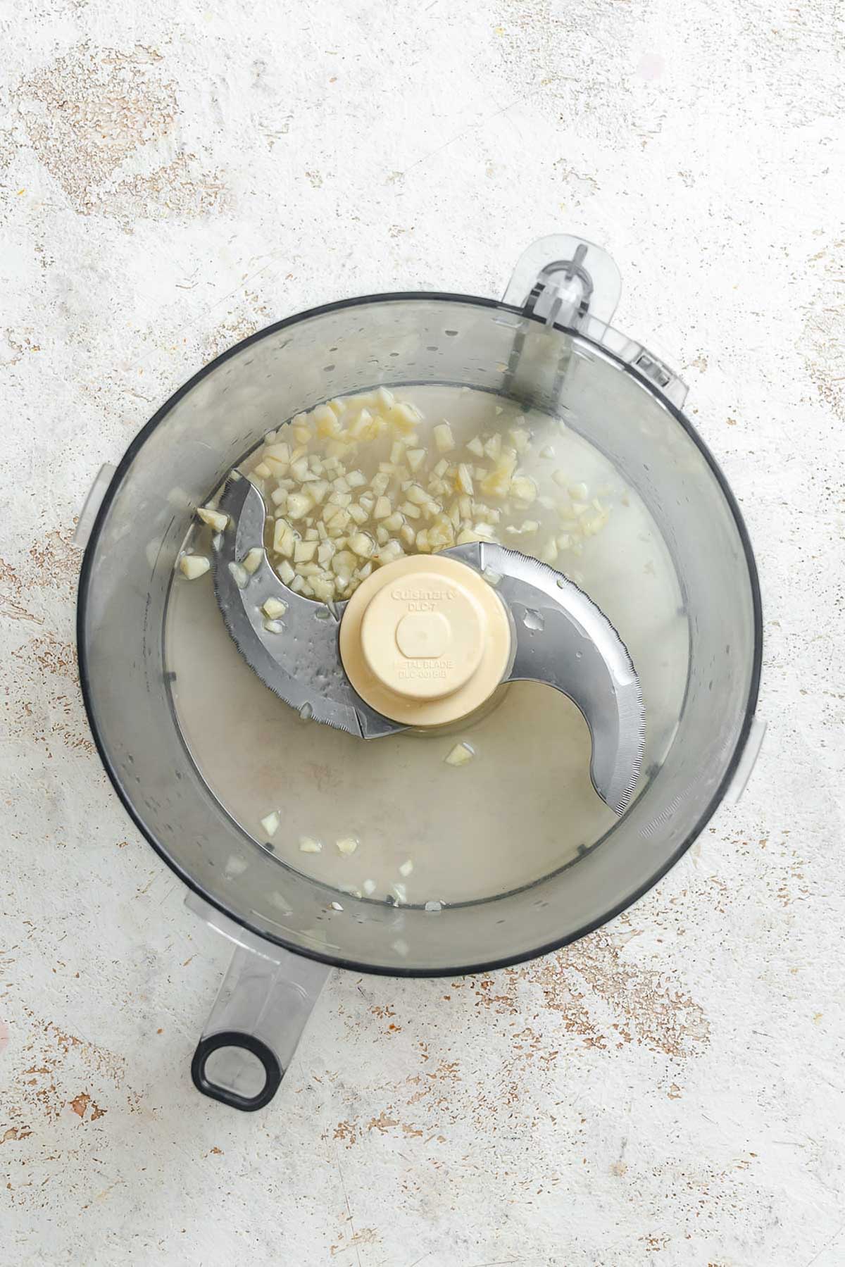 Chopped raw garlic pieces sitting in lemon juice at the bottom of a food processor bowl.