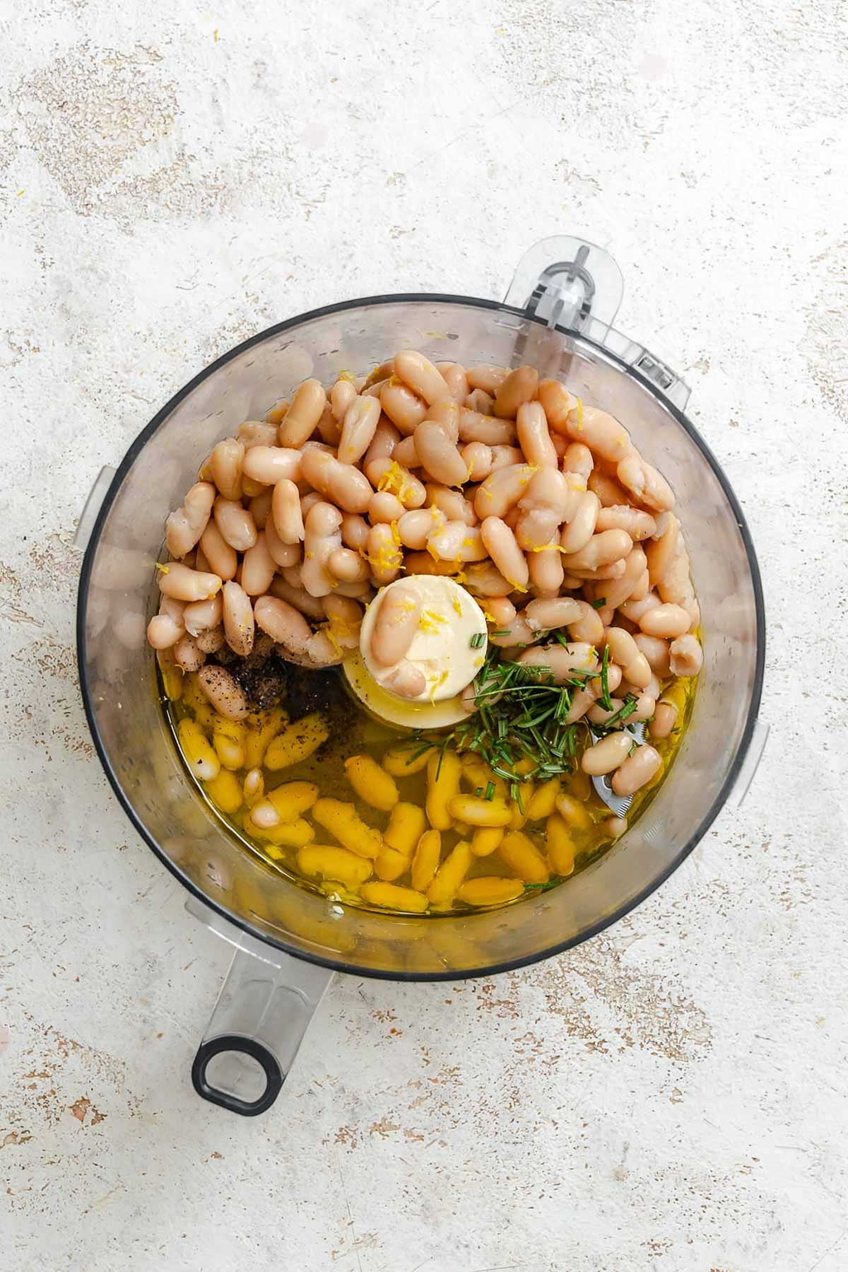Cannellini beans, olive oil, lemon zest, cracked black pepper, and fresh rosemary sprigs added to a food processor before blending.