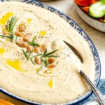 Creamy white bean dip in a blue patterned serving bowl, topped with whole beans, rosemary, black pepper, and a drizzle of olive oil, with fresh vegetables on the side.