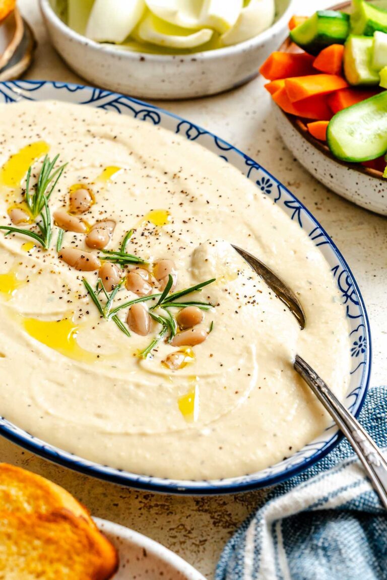 Creamy white bean dip in a blue patterned serving bowl, topped with whole beans, rosemary, black pepper, and a drizzle of olive oil, with fresh vegetables on the side.