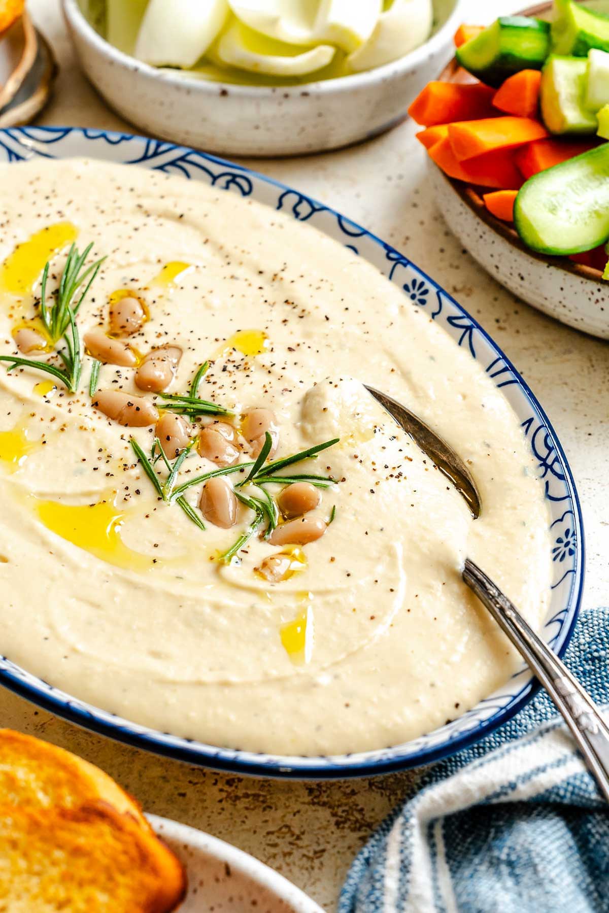 Creamy white bean dip in a blue patterned serving bowl, topped with whole beans, rosemary, black pepper, and a drizzle of olive oil, with fresh vegetables on the side.