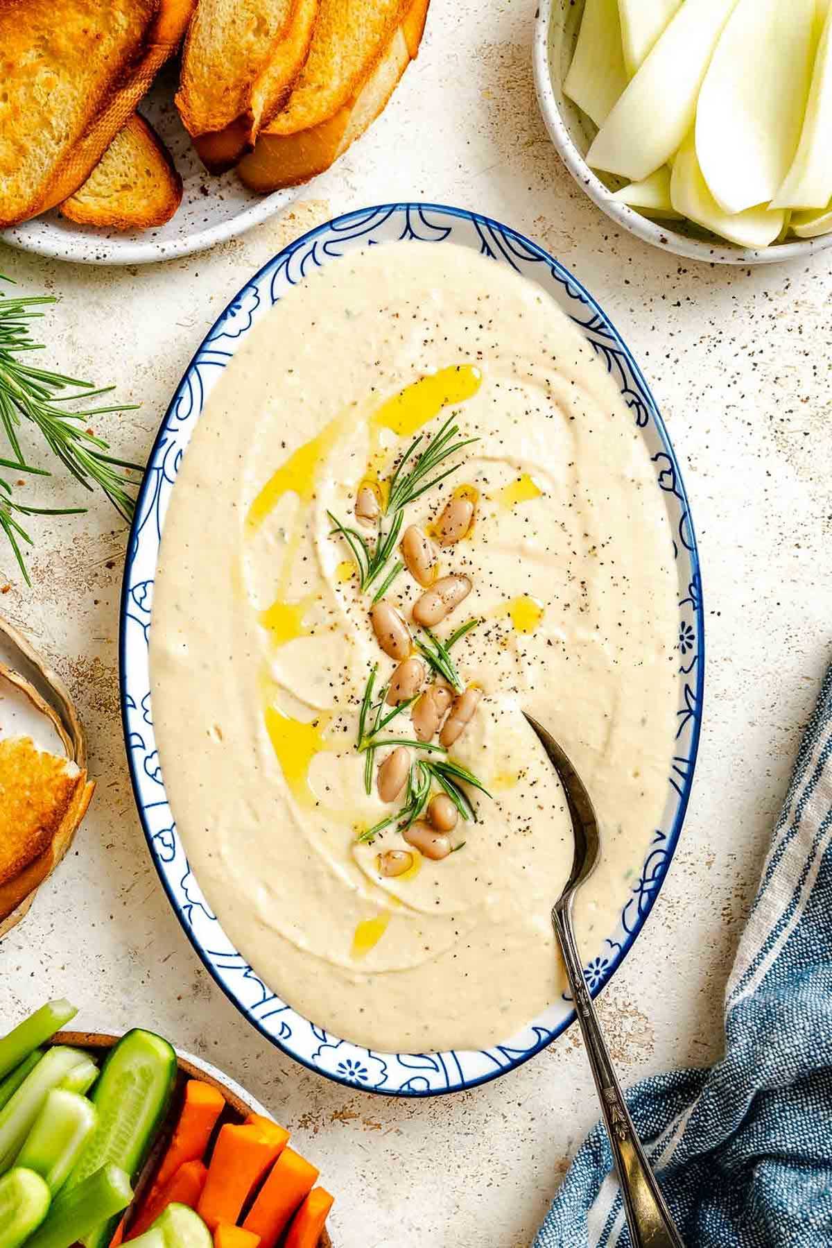 Overhead view of creamy white bean dip in a blue patterned bowl, garnished with rosemary and olive oil, surrounded by toasted bread and cut vegetables.