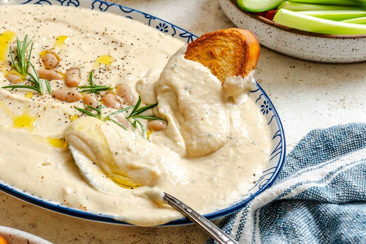 Creamy white bean dip served in a shallow bowl, with toasted bread dipped in, showing its thick, smooth consistency.