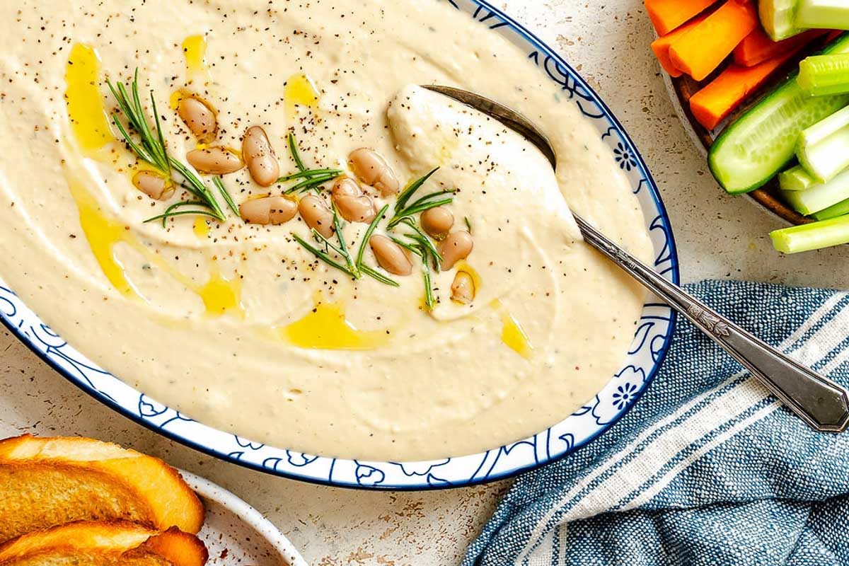 Overhead view of the dip garnished with rosemary, olive oil, and beans, served with carrot sticks, cucumber slices, and toasted bread.