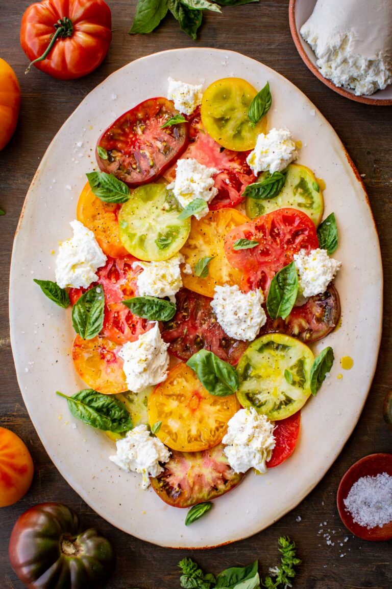 Overhead shot of completed Caprese salad on a platter, surrounded by fresh tomatoes, basil, and a bowl of flaky salt.
