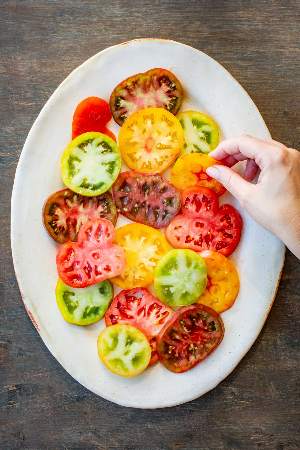 Sliced heirloom tomatoes in shades of red, yellow, and green arranged on a white serving platter with a hand placing one slice.