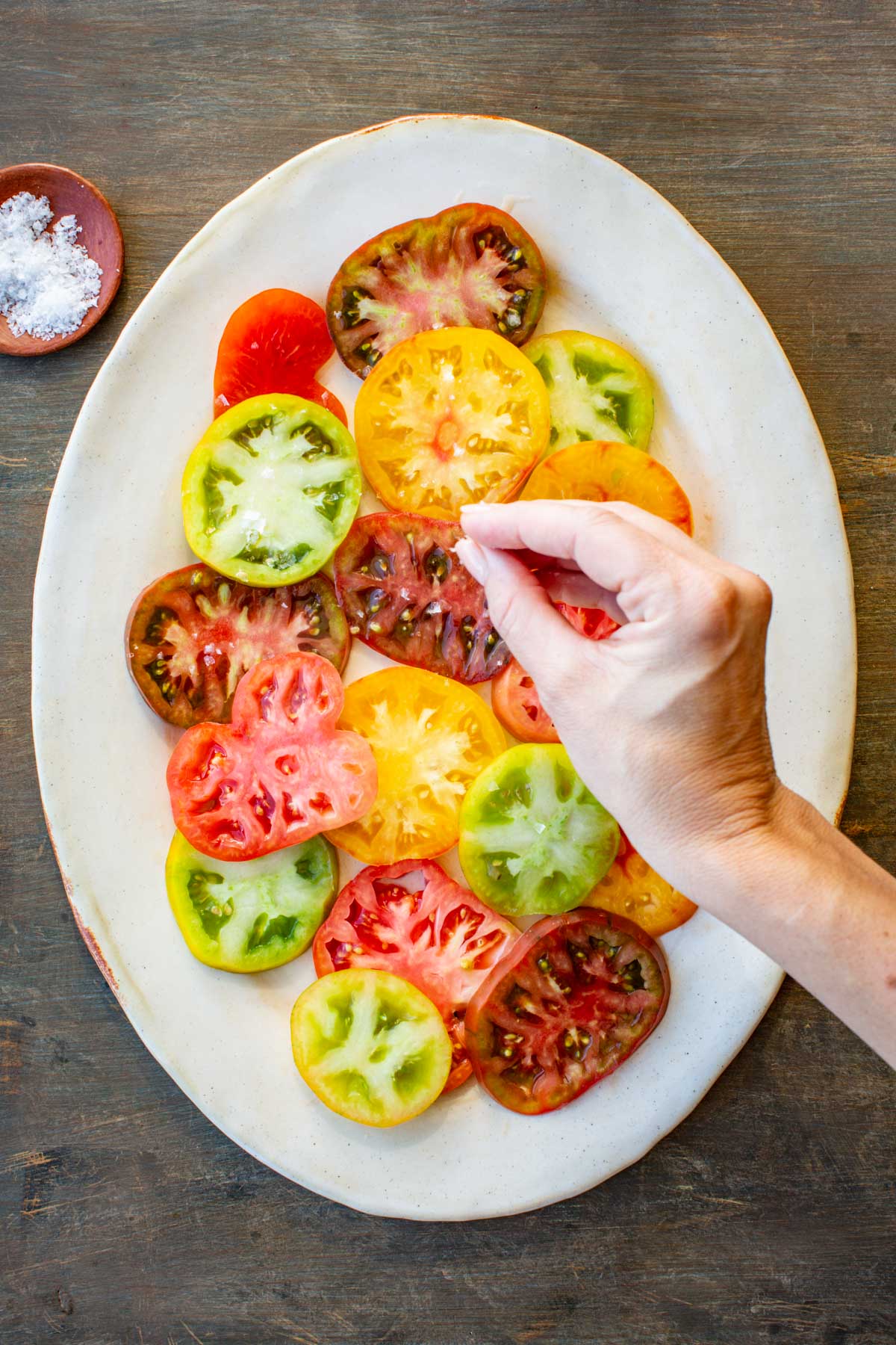 Hand sprinkling flaky sea salt over colorful tomato slices on a platter with a small bowl of salt nearby.