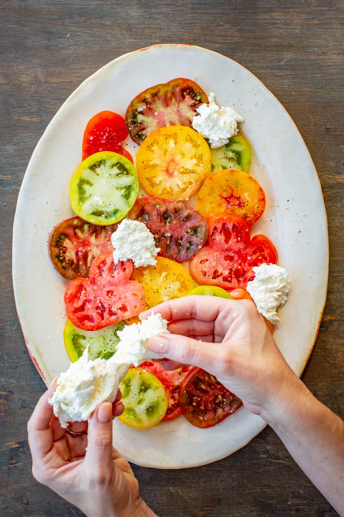 Hand tearing fresh mozzarella and placing rustic chunks over seasoned tomato slices on a serving plate.