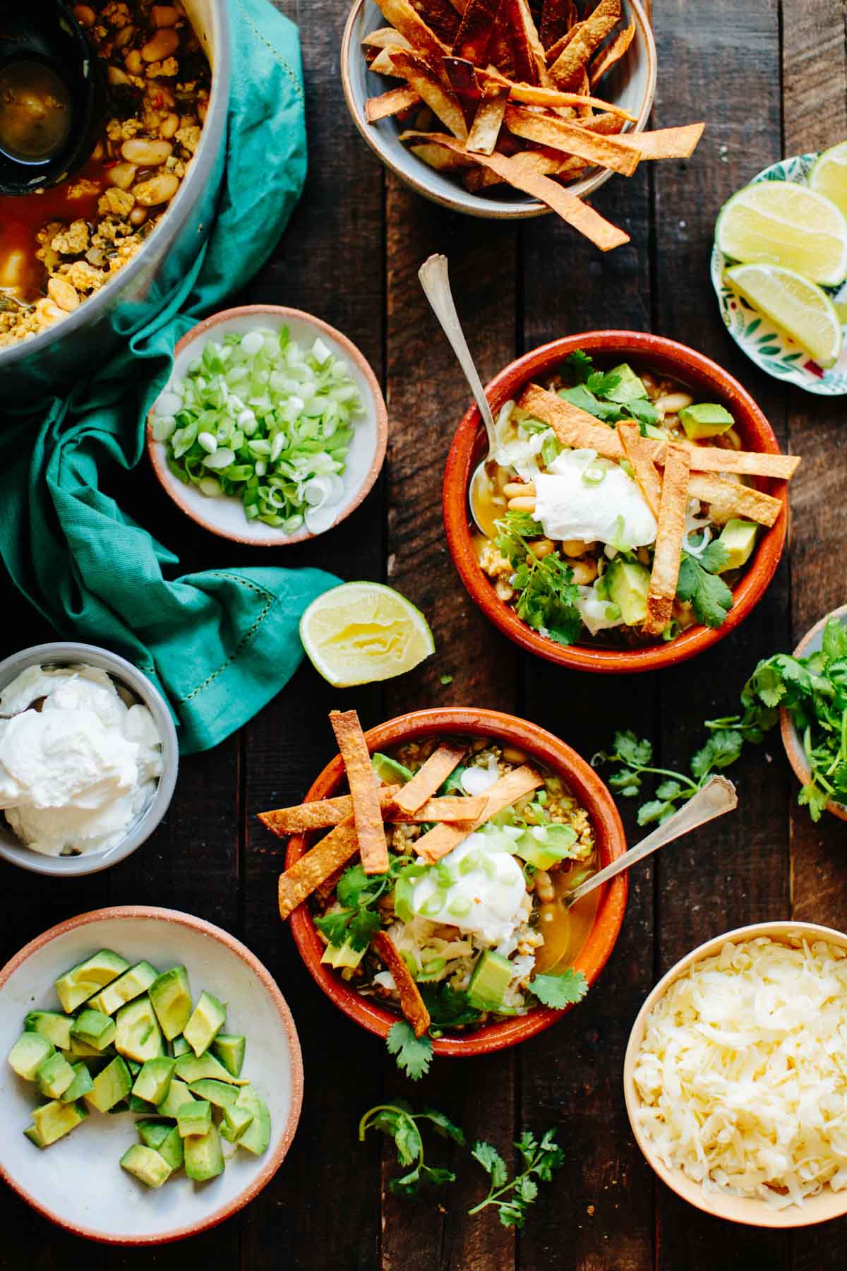 Bowls of creamy white chicken chili surrounded by toppings like avocado, shredded cheddar cheese, lime wedges, sour cream, scallions, cilantro, and crispy tortilla chips.