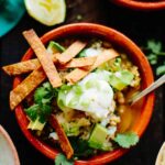 A close-up of a clay bowl of chili with a creamy texture, topped with sour cream, avocado chunks, fresh cilantro, scallions, and crunchy tortilla strips.