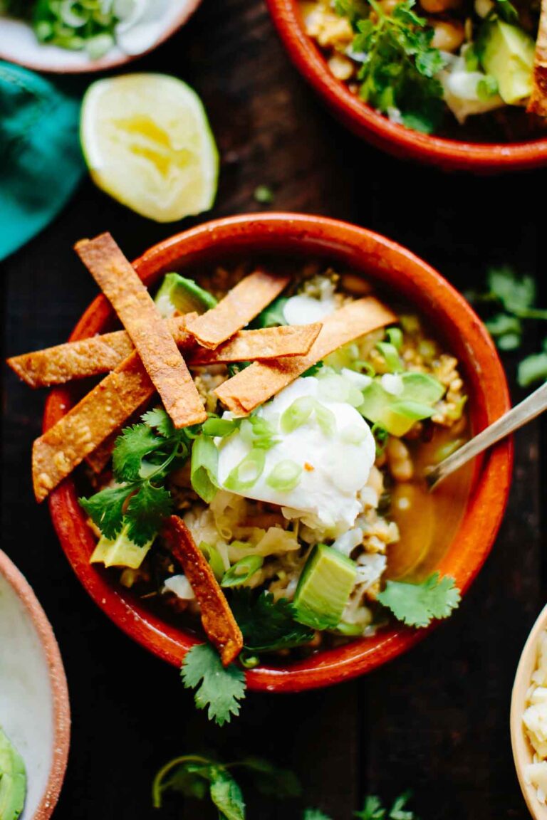 A close-up of a clay bowl of chili with a creamy texture, topped with sour cream, avocado chunks, fresh cilantro, scallions, and crunchy tortilla strips.