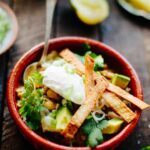 A single serving of white chicken chili in a red clay bowl garnished with green onions, avocado, tortilla chips, and a dollop of sour cream.