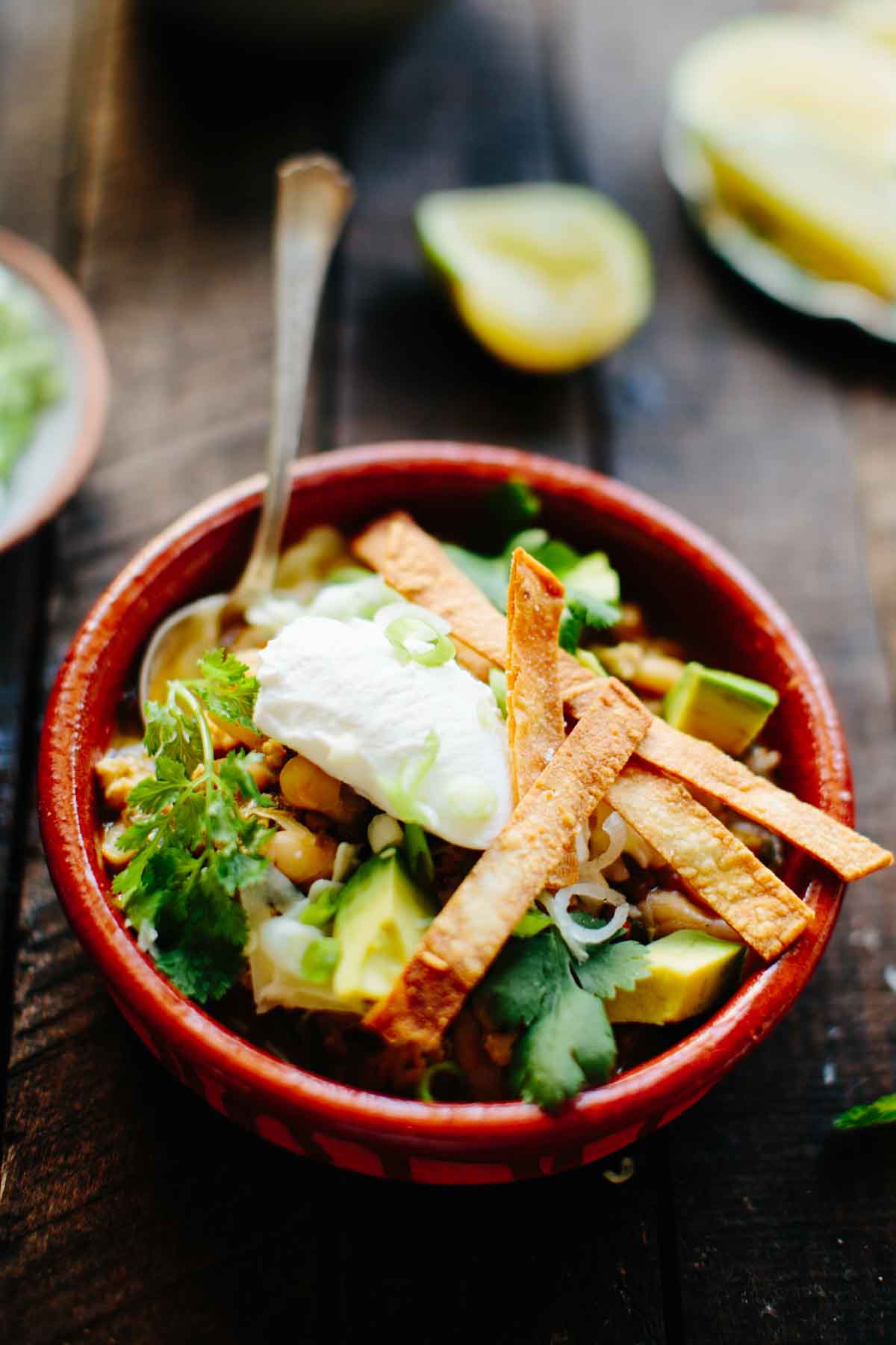 A single serving of white chicken chili in a red clay bowl garnished with green onions, avocado, tortilla chips, and a dollop of sour cream.