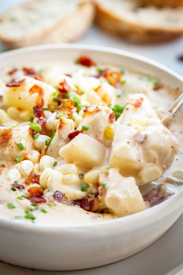 Close-up of a spoonful of creamy chowder lifted from a bowl, showing chunks of potato, sweet corn, bacon, and crab meat in a rich broth with bread in the background.