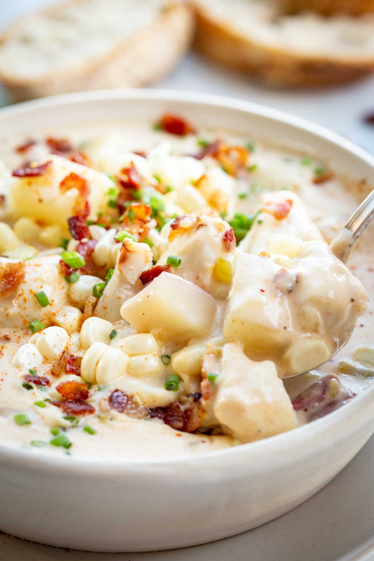 Close-up of a spoonful of creamy chowder lifted from a bowl, showing chunks of potato, sweet corn, bacon, and crab meat in a rich broth with bread in the background.