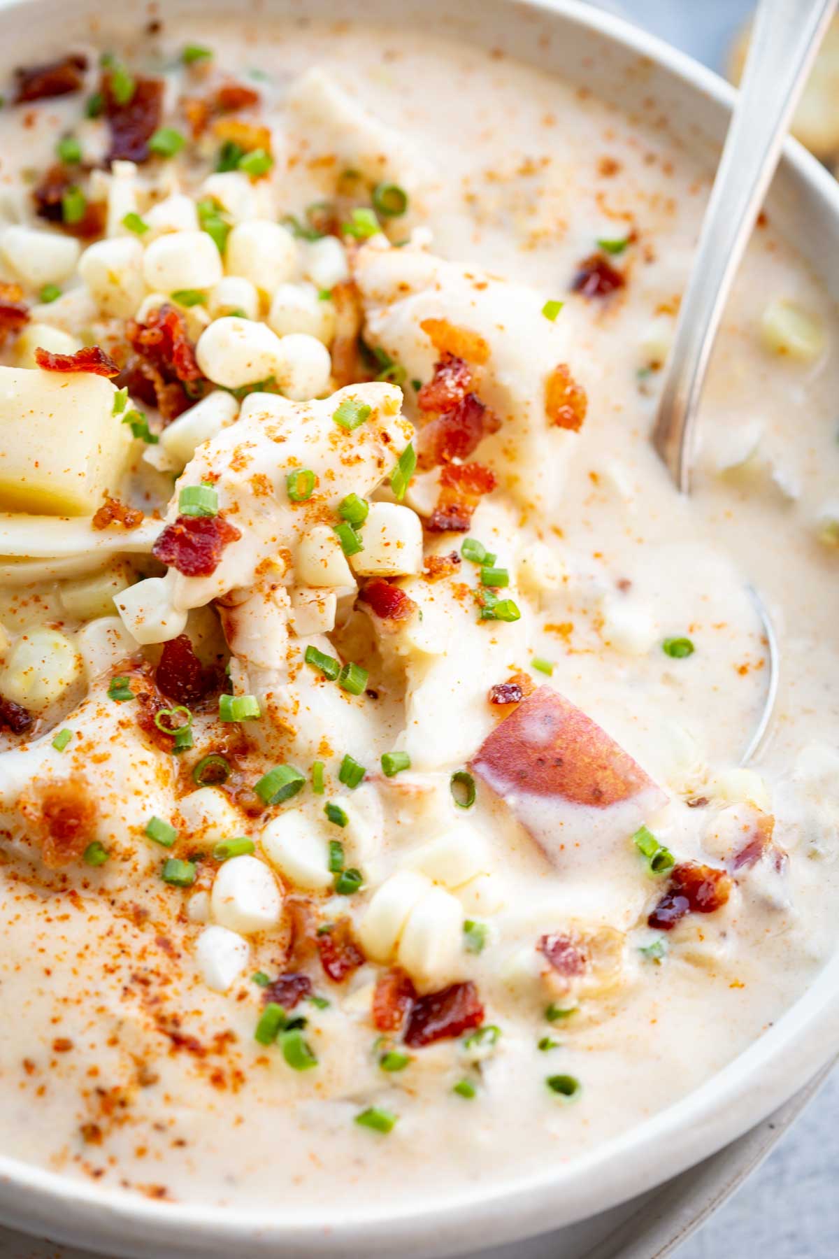 A spoon resting in a bowl of chowder filled with potatoes, corn, bacon, and crab meat, all in a thick, creamy broth topped with fresh chives.