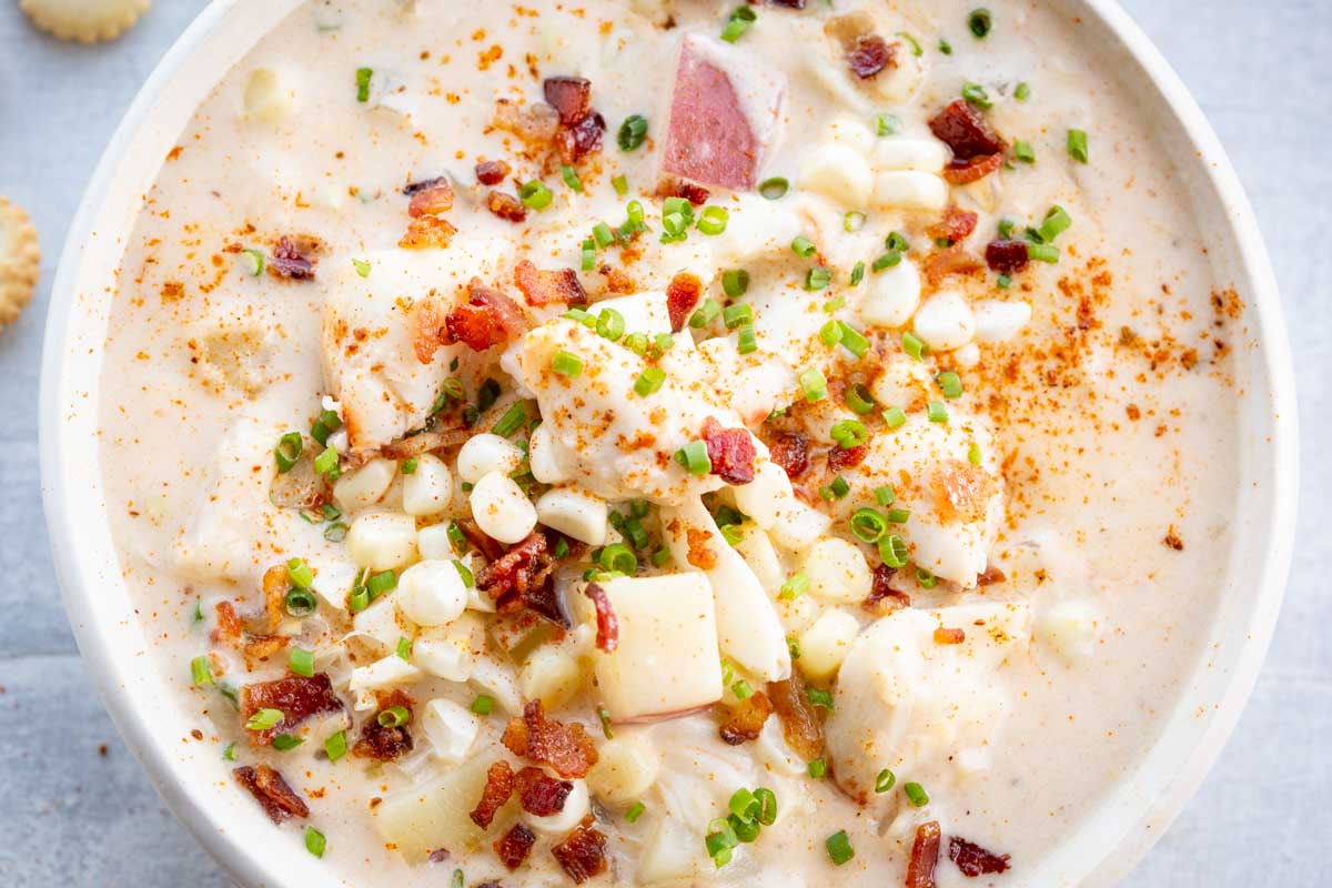 Overhead view of a bowl of corn and crab chowder showing large chunks of potato, corn, and crab meat garnished with bacon and chives, dusted with seasoning.