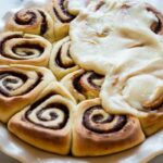 Freshly baked cinnamon rolls in a white dish, with rich cream cheese frosting spread over half, contrasting with the unfrosted swirls.