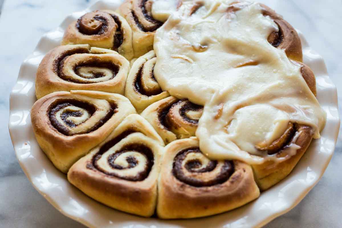 Freshly baked cinnamon rolls in a white dish, with rich cream cheese frosting spread over half, contrasting with the unfrosted swirls.