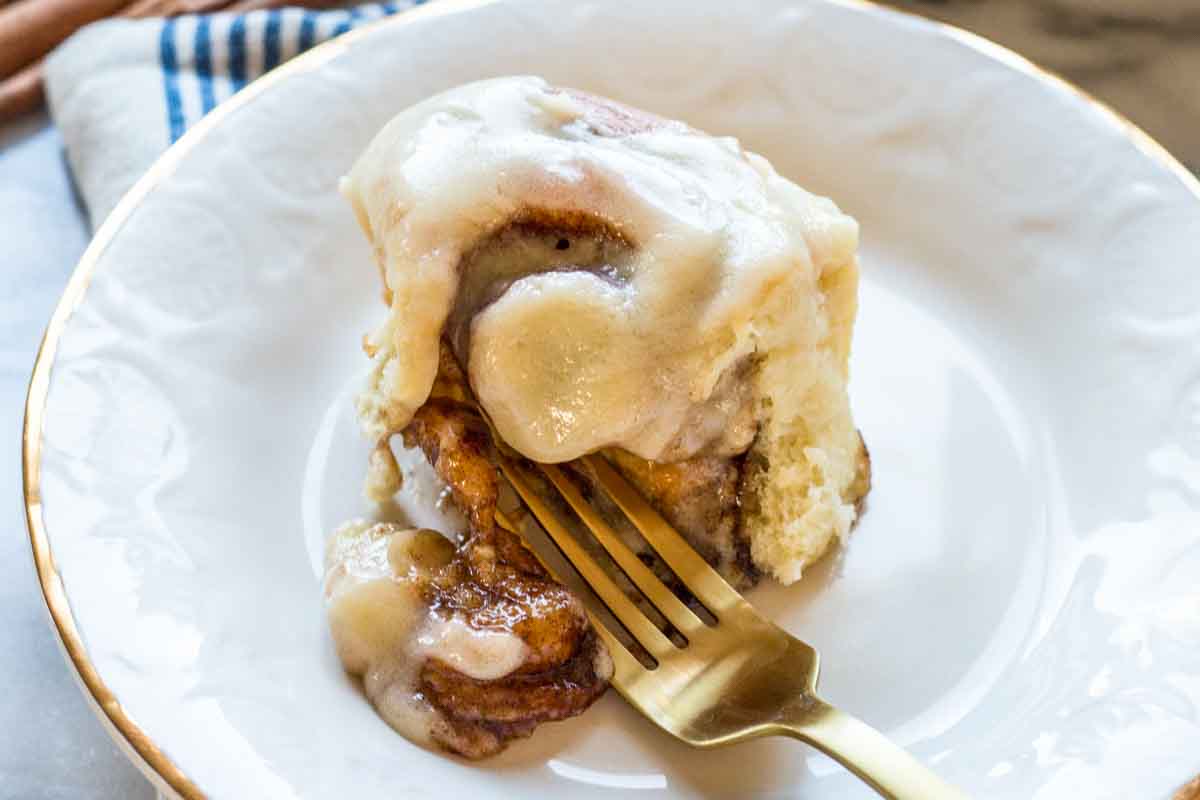 A fluffy cinnamon roll on a white plate, generously coated in cream cheese frosting with a golden fork cutting into it.