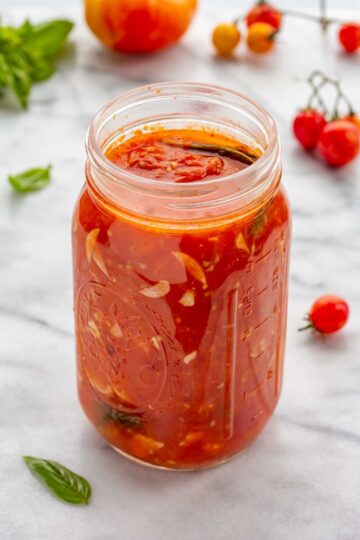 Mason jar filled with chunky homemade tomato sauce on a marble surface, surrounded by fresh tomatoes and basil leaves.