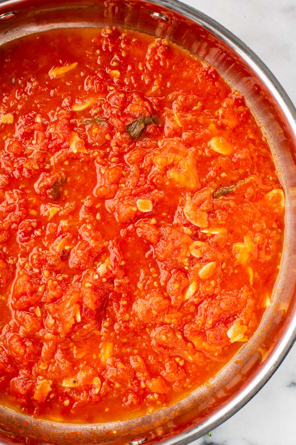 Close-up of fresh tomato sauce simmering in a stainless steel pan, with garlic slices and basil visible in the sauce.
