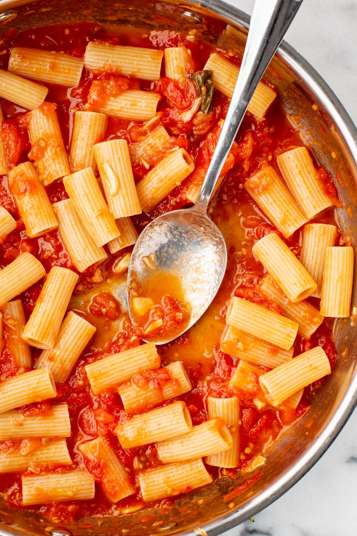 Rigatoni pasta stirred into the tomato sauce in a pan with a large silver spoon.