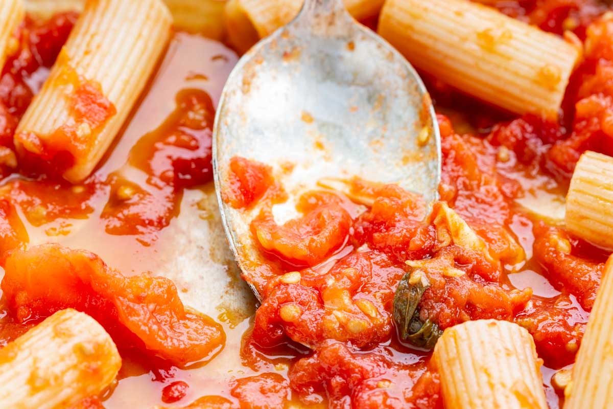 Close-up of spoon holding a scoop of tomato sauce with rigatoni pasta scattered around in the pan.
