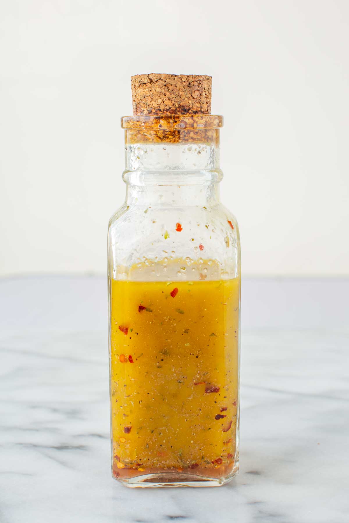 A glass bottle filled with freshly shaken Italian salad dressing, golden yellow with visible flecks of red pepper flakes and black pepper.