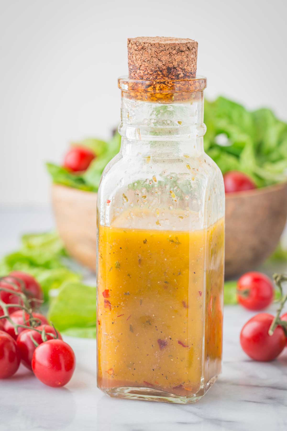 Close-up of a glass bottle of zesty Italian dressing surrounded by salad greens, basil, and cherry tomatoes on the table.