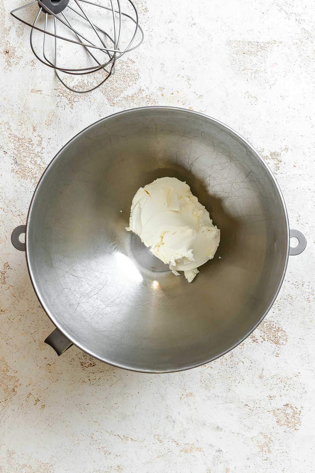 Mascarpone cheese placed in the bottom of a large stainless steel mixing bowl with a whisk attachment nearby.